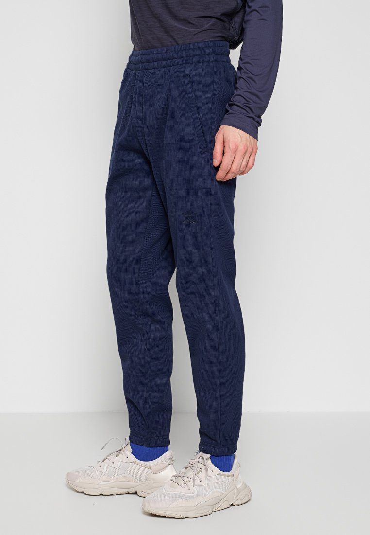 Marin blauwe joggingsbroek met textuur, elastische tailleband, zijzakken en geborduurd logo. Gecombineerd met lichtbeige sneakers en blauwe sokken.