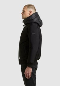Czarna puchowa kurtka z wysokim, wyściełanym kołnierzem i zapinana na zamek błyskawiczny z przodu. Na rękawie znajduje się materiałowa naszywka, a rękawy zakończone są elastycznymi ściągaczami. Powierzchnia o fakturze.