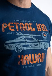 Marineblå bomulds T-shirt med et grafisk tryk af en bil med surfbrætter; orange og grå tekst med teksten "Petrol Ind." og "Hawaii." Glat tekstur.