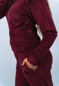 Bordeauxkleurige top met ritssluiting en broek van zachte, gestructureerde stof; heeft een zak met zwarte knoopdetail en nauwsluitende mouwen.