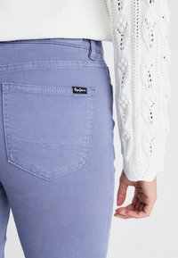 Violetinės džinsinės kelnytes su prigludusia forma, turinčios siuvinėtą užpakalinį kišenę ir juodą Pepe Jeans logotipo etiketę. Tekstūra atrodo lygi su nedideliu blizgesiu.