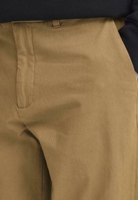 Kāhkola krāsas bikses no kokvilnas maisījuma auduma ar gludu faktūru, taisnu piegriezumu un sānu kabatām. Redzami vienkārši metāla detaļas.