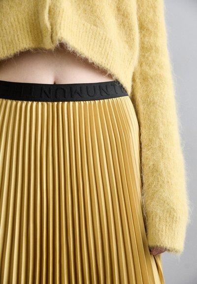 Pulover tricotat galben și pufos, asortat cu o fustă plisată galben-mustar, care prezintă un brâu elastic negru cu inscripții embosate.
