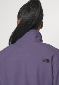 Fioletowa polarowa kurtka z wysokim kołnierzem i teksturowanym wykończeniem. Czarny logo na górnej lewej części pleców. Brak widocznych zapięć lub wzorów.