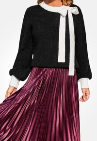 Maglione nero lavorato a maglia con dettaglio in nastro bianco e maniche lunghe, abbinato a una gonna plissettata bordeaux, che evidenzia una texture lucida.