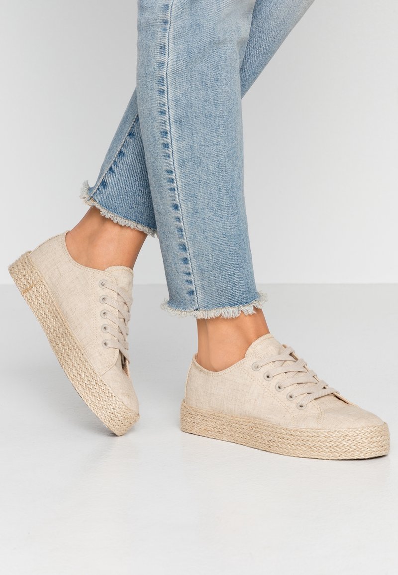 Beige kanvas sneakers dengan sol espadrille yang tekstur, desain tali, dan jeans biru dengan tepi yang sobek.
