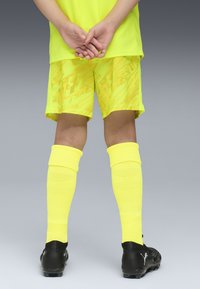 Camiseta y pantalones cortos deportivos amarillos con patrones abstractos, combinados con calcetines amarillos hasta la rodilla y botines de fútbol negros, vistos desde atrás.