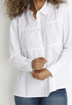 Femme aux cheveux bouclés portant un chemisier blanc boutonné et un jean bleu, ajustant le poignet de sa manche gauche avec sa main droite.