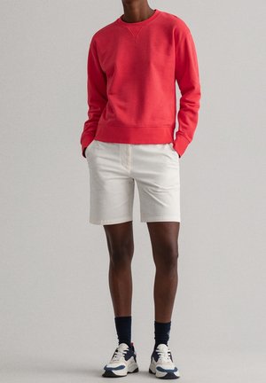 Osoba ubrana w jaskrawą czerwoną bluzę z długim rękawem, kremowe szorty do kolan, ciemne skarpetki oraz białe trampki z niebieskimi akcentami, stojąca z rękami w kieszeniach.