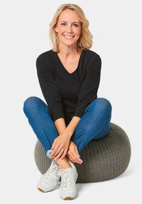 Grijze gebreide pouf met gestructureerd oppervlak, vergezeld door een persoon in een zwart langmouwenshirt en blauwe spijkerbroek, casual zittend.