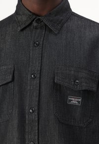 Black denim shirt featuring a button-up front, two chest pockets, and a label on the left pocket. Textured fabric with a slight sheen.