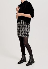 Femme portant un sweat à capuche noir, une jupe à motif pied-de-poule, des collants noirs et des bottines noires à lacets, debout devant un fond uni.