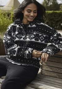 Svart och vit fleece-pullover med geometriska mönster, halvdragkedja och långa ärmar, kombinerad med svarta sweatpants.