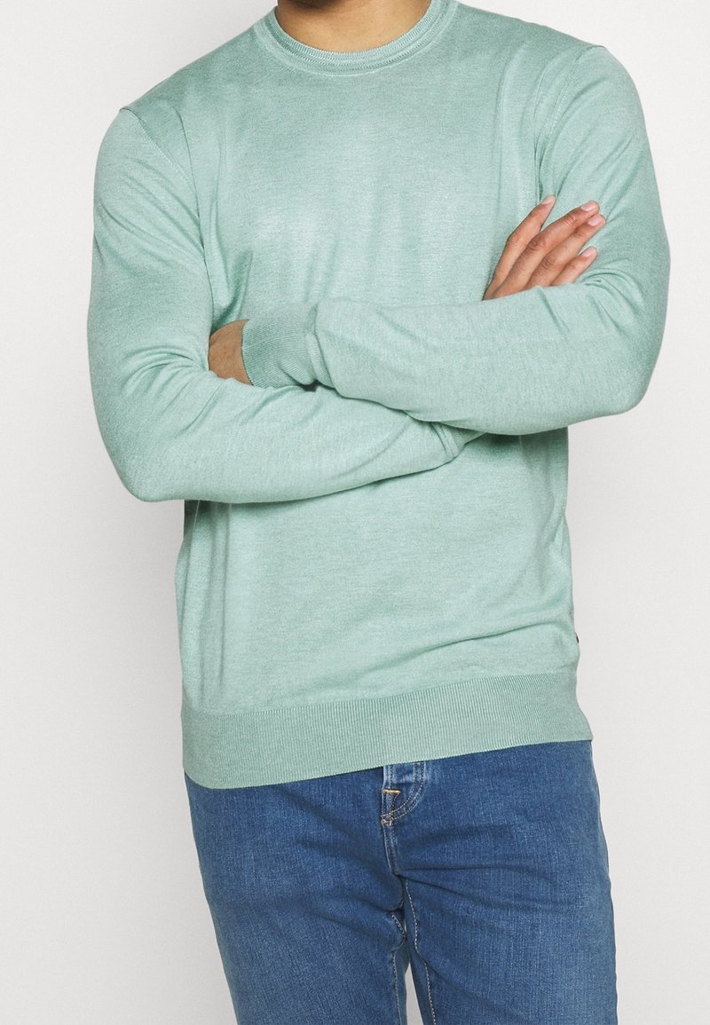 Personne portant un pull à manches longues vert clair, bras croisés, associé à un jean bleu en denim, debout devant un fond uni.