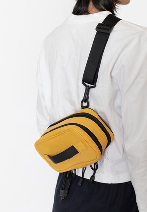 Borsa sintetica gialla con tre scomparti con cerniera, dettagli neri e tracolla regolabile, indossata diagonalmente sul corpo.