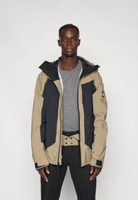 Μπεζ και ναυτικό μπλοκαρισμένο μπουφάν με κουκούλα, με φερμουάρ μπροστά, ελαστικές μανσέτες και ρυθμιζόμενο τελείωμα. Φορεμένο πάνω από γκρι πουκάμισο.