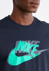 Detailní záběr osoby v černém tričku s vrstveným tyrkysovým a šedým logem Nike na hrudi.