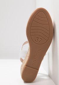 Valkoinen sandaali, jossa on espadrille-tyylinen pohja. Karkea beige ulkopinta, sileät synteettiset hihnat ja kuminen ruskea pohja, jossa on merkintä "37".