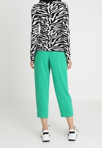 Osoba ubrana w czarną i białą bluzkę w zebrę z długim rękawem, zielone krótkie spodnie oraz białe sneakersy, stojąca na tle jednolitym.