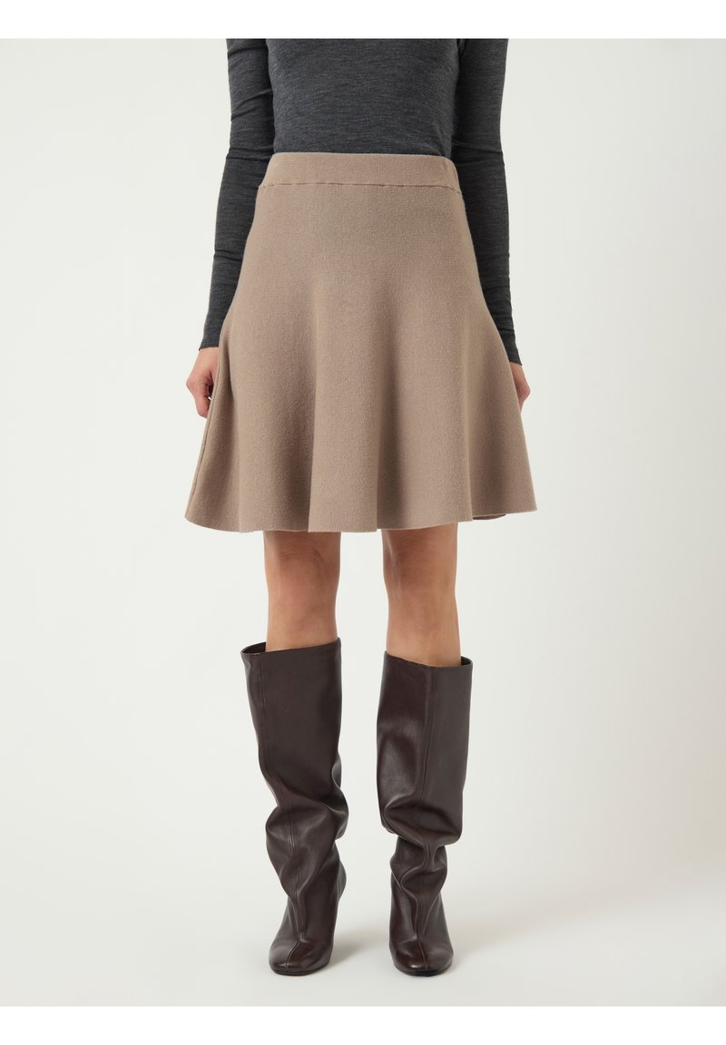 Jupe beige longueur genou en forme trapèze portée avec un haut à manches longues gris foncé et des bottes en cuir marron foncé, souples, montant jusqu'aux genoux, sur un fond uni.