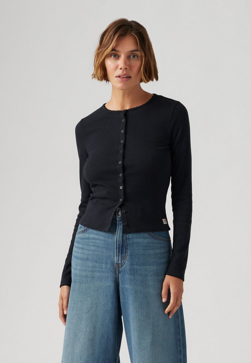 Cardigan noir à manches longues en tissu côtelé, avec un col rond et une fermeture à boutons à l'avant, associé à un jean taille haute bleu clair à jambe large.