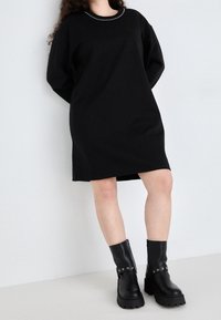Robe noire surdimensionnée avec un col rond et des clous décoratifs en argent, associée à des bottines noires à bout arrondi avec une sangle et du matériel en argent.