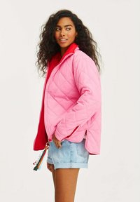 Prešívaná ružová bunda s červenou vnútornou podšívkou, obsahujúca zips, bočné rozparky a farebný strapec na ťahadle zipsu.