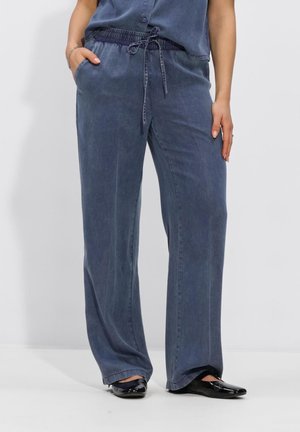 Femme portant un pantalon ample bleu foncé à taille élastiquée avec cordon, mains dans les poches, associé à des chaussures plates noires brillantes.