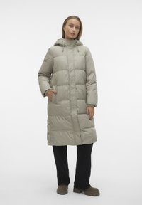 Lang beige pufferjas met capuchon, verstelbare trekkoorden, voorzakken en knoopsideopeningen, gedragen over donkere broek en schoenen.