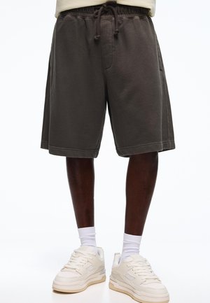 Tmavomodré šortky so šnúrkou nosené s bielymi ponožkami a béžovými teniskami na osobe stojacej na bielom pozadí.