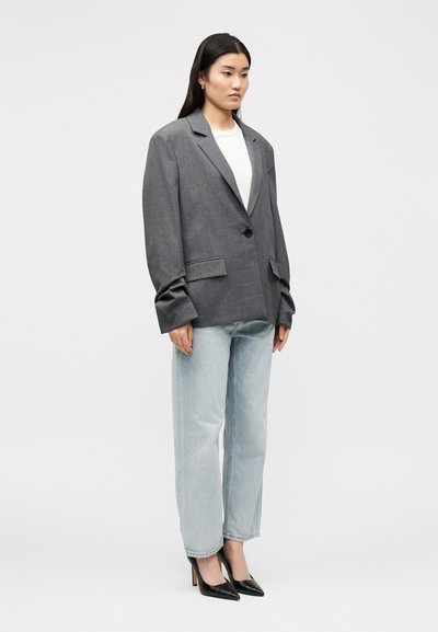 Γυναίκα που στέκεται φορώντας μια φαρδιά γκρι σακάκι, λευκό τοπ, ανοιχτό μπλε τζιν και μαύρες μυτερές ψηλοτάκουνες γόβες, πάνω σε απλό ανοιχτό φόντο.