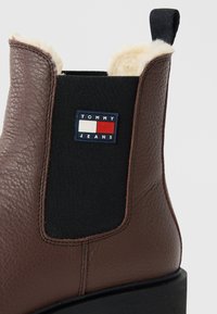 Brune lærstøvle med sort elastikpanel, hvid fleeceforing, trækstrop og Tommy Jeans-logo patch.
