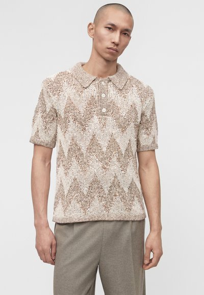 Άνδρας που φοράει ένα πλεκτό πόλο με κοντά μανίκια και σχέδιο ζιγκ-ζαγκ σε μπεζ και λευκό χρώμα, με ανοιχτό γκρι παντελόνι, σε απλό φόντο.