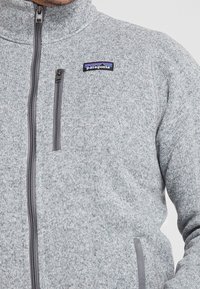 Grå fleece zip-up jacka med hög krage, som har en logotyp-patch och en kontrasterande grå dragkedja. Materialet har en strukturerad, stickad look.
