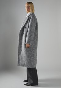 Langer grauer Wollmantel mit strukturiertem Finish, doppelten Knöpfen und Seitentaschen. Kombiniert mit schwarzen Schlaghosen und spitzen schwarzen Schuhen.