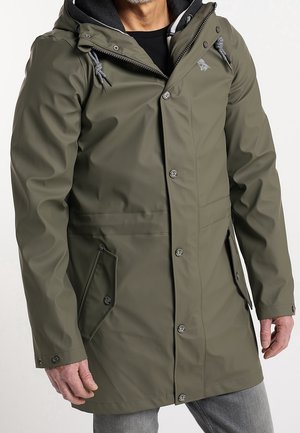 Homme portant un imperméable à capuche vert olive avec boutons devant, zip, cordons et poches à rabat, debout devant un fond uni.