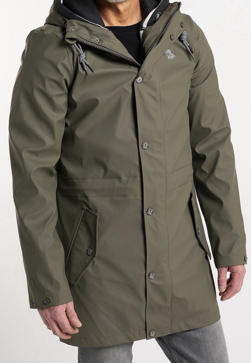 Homme portant un imperméable à capuche vert olive avec boutons devant, zip, cordons et poches à rabat, debout devant un fond uni.