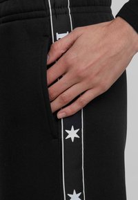 Črne trenirke imajo stransko žep in bele zvezdaste vzorce ob strani, ter bele obrobe. Roka počiva na območju žepa.