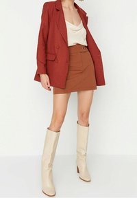 Femme portant un blazer rouille, un haut beige, une jupe assortie rouille et des bottes à talons hautes crème jusqu'aux genoux, posant devant un fond blanc.