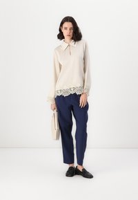 Blusa crema con pizzo sull'orlo, abbinata a pantaloni navy plissettati. La modella tiene una borsa beige e indossa mocassini neri.