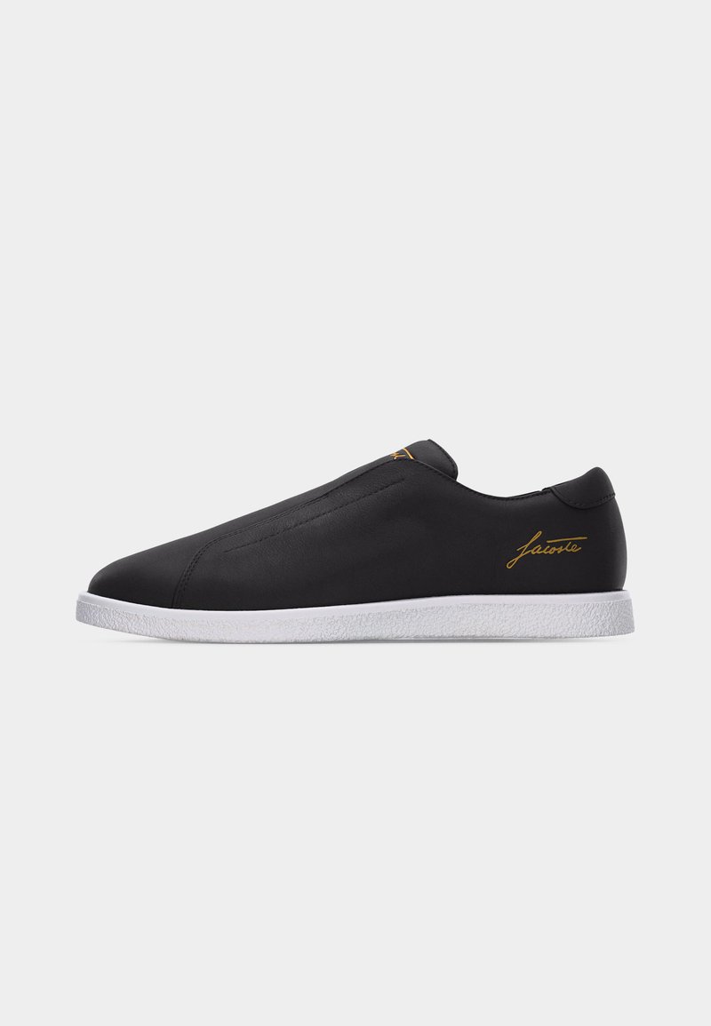 Sneaker nero slip-on con suola bianca e firma dorata "Lacoste" sul tallone e sulla linguetta, vista di lato su sfondo bianco.