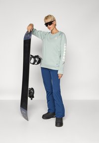 Sudadera verde menta con detalle del logo en blanco, pantalones de nieve azules, botas de snowboard negras y una tabla de snowboard negra con fijaciones.