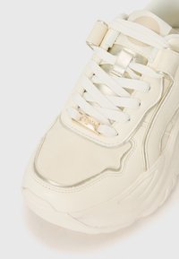 White sneaker with textured material, metallic accent on the side, and gold branding. Features white laces and a padded collar for comfort.