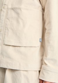 Dettaglio della tasca della giacca di velluto a coste beige con chiusura a bottone e piccola etichetta di tessuto grigia del marchio sul polsino della manica.