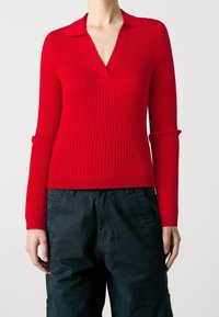 Roter, gerippter Strickpullover mit V-Ausschnitt und spitzem Kragen. Lange Ärmel mit ausgeschnittenen Akzenten. Kombiniert mit dunklen Cargo-Hosen.