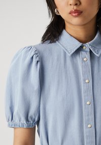 Ανοιχτό μπλε τζιν πουκάμισο με κλασικό γιακά, κοντά φουσκωτά μανίκια με ελαστικές μανσέτες και μπροστινό κλείσιμο με κουμπιά από μέταλλο.