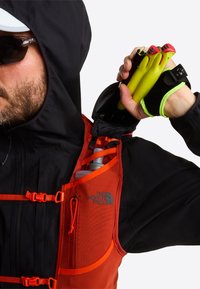 Pomarańczowa kamizelka z siateczkowego materiału z czarnymi akcentami, wyposażona w zamki błyskawiczne, regulowane paski oraz kieszeń na butelkę z wodą. Zielone kijki trzymane w ręku.