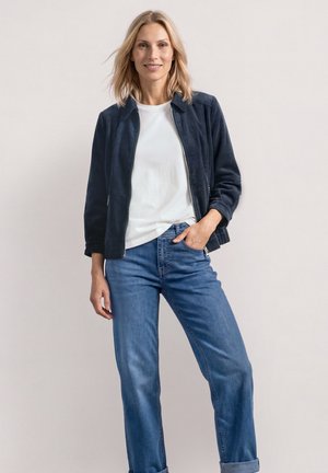 Frau mit blondem Haar, die eine dunkelblaue Jacke, ein weißes T-Shirt und blaue Jeans trägt, steht mit einer Hand in der Tasche vor einem einfarbigen Hintergrund.