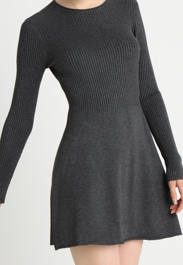 Femme portant une robe en maille gris foncé à manches longues avec un haut côtelé et une jupe évasée lisse, debout devant un fond uni.