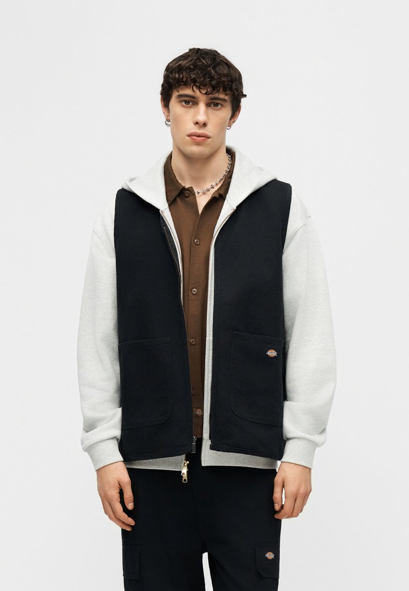 Giovane uomo con capelli corti e ricci che indossa un gilet nero sopra una felpa grigia con cappuccio e una camicia marrone con bottoni, in piedi davanti a uno sfondo bianco uniforme.